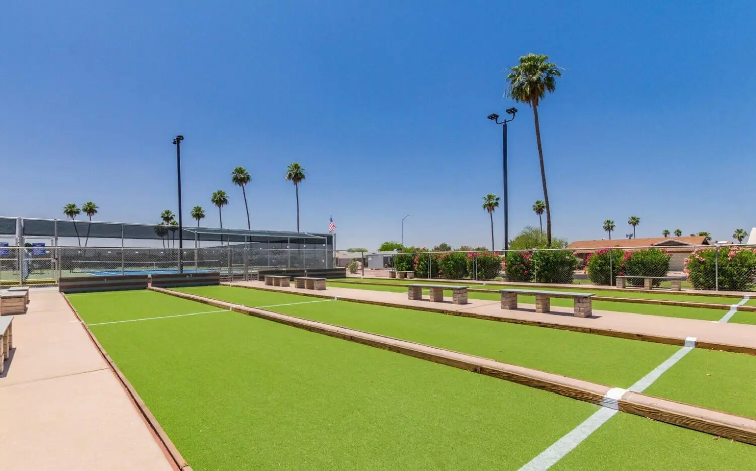 A sunny outdoor sports area in Deer Valley features two bocce ball courts lined with lush artificial grass. Tall palm trees and trimmed bushes surround the courts under a clear blue sky. Benches are placed along the side of the courts for relaxation.