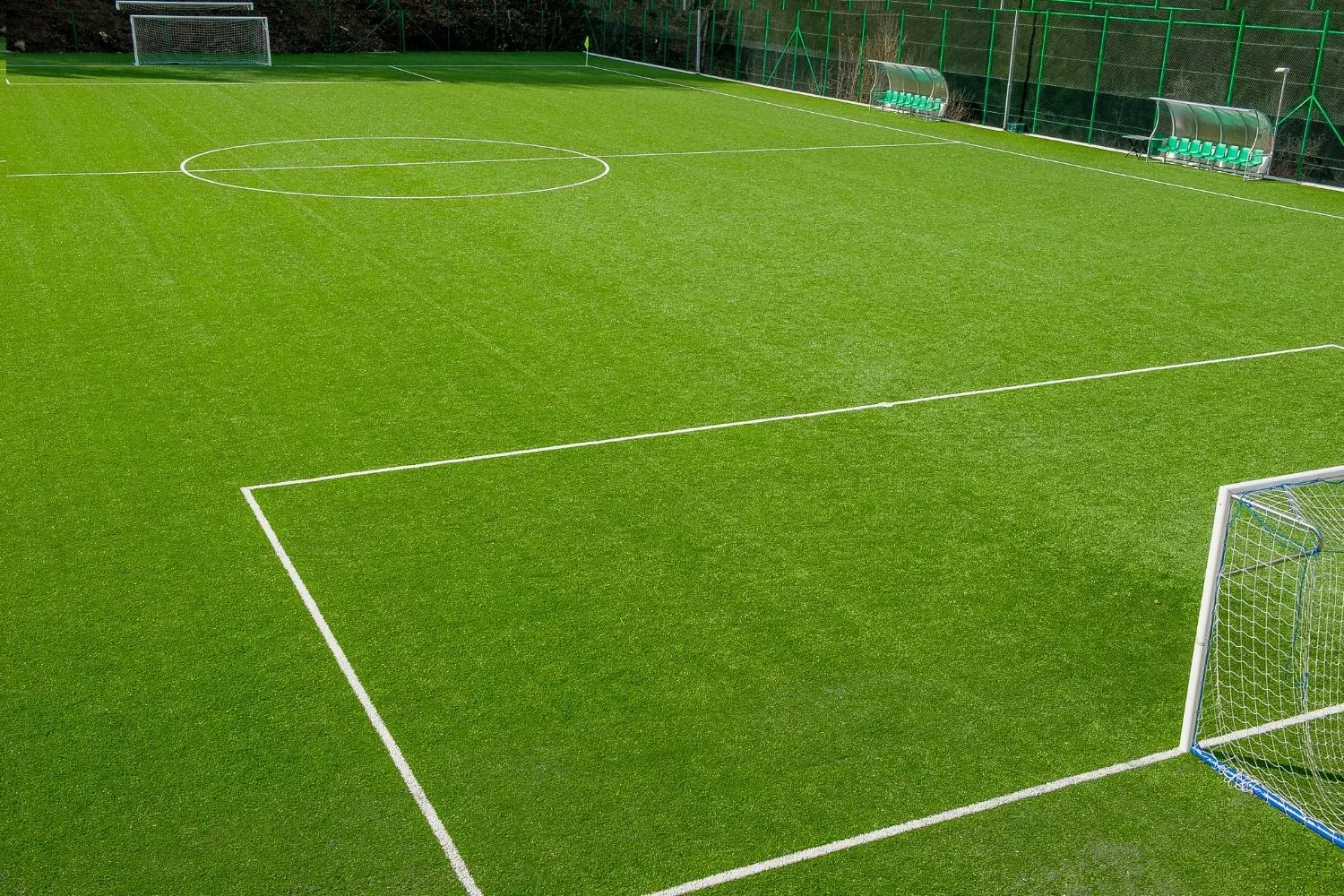 The Deer Valley green soccer field, featuring pristine white boundary lines, showcases two goals—one in the foreground and another in the background. Surrounded by a green fence and visible team benches, it's a perfect example of quality synthetic turf installation offering top-notch artificial grass solutions.