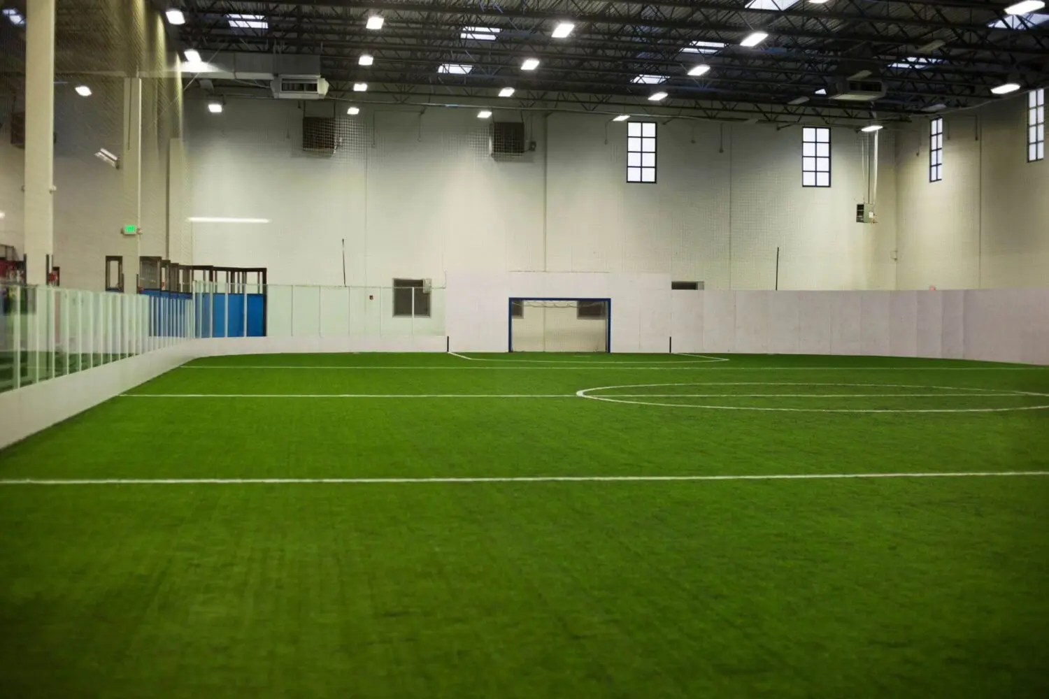 The indoor soccer field boasts synthetic turf installation with white walls. Marked lines and a goalpost feature prominently against the vivid greenery. Large windows and overhead lights illuminate the space, while glass partitions separate it from the viewing area in Deer Valley North Phoenix.