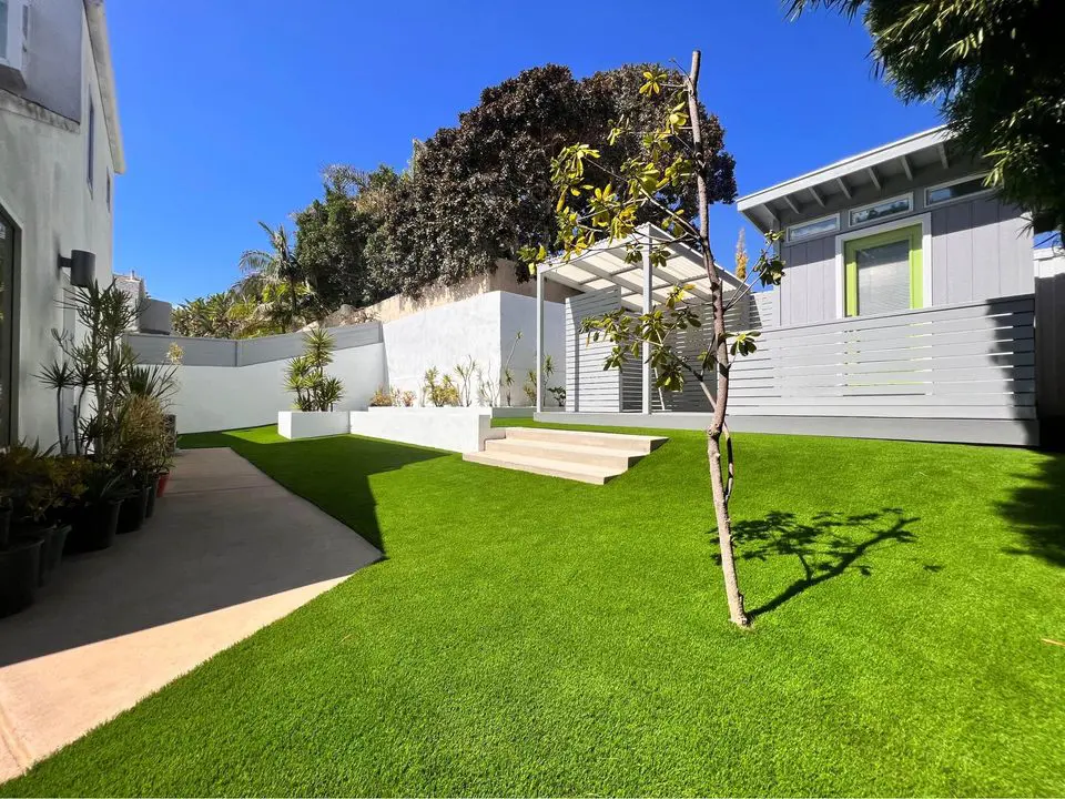 A modern backyard in Deer Valley, Arizona, features a small tree, manicured lawn, and a paved path. The contemporary gray-and-white shed stands out against the artificial grass installations, surrounded by a white fence and lush greenery under the clear blue sky.