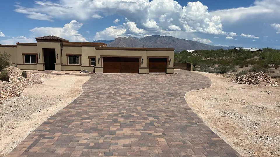 A modern, single-story desert home boasts exceptional curb appeal with a long, elegantly paved driveway. The house features a flat roof and a two-car garage. In the background, mountains and a partly cloudy sky enhance the serene setting, while sparse desert vegetation completes this Deer Valley gem.