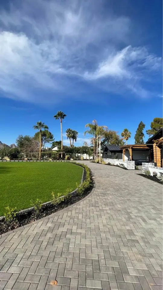 A brick pathway with undeniable curb appeal curves through a lush, green lawn bordered by palm trees. A modern house with a wooden facade is seen on the right. The sky is clear with a few wispy clouds, creating a serene and inviting atmosphere reminiscent of Deer Valley.