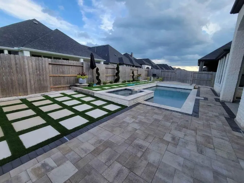 A modern backyard in Deer Valley, AZ, boasts a rectangular swimming pool with an adjacent hot tub. The area showcases curb appeal with large tile paver installations and patches of artificial grass. Decorative sculptures and potted plants add charm under a cloudy sky.