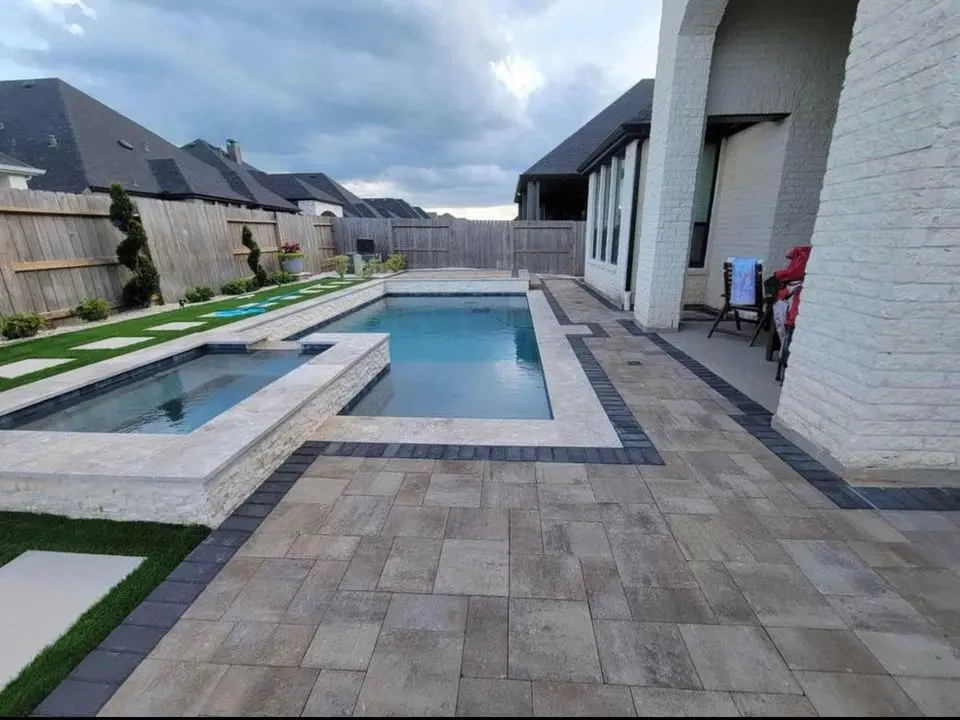 A modern backyard in Deer Valley boasts a large rectangular pool and a smaller adjacent hot tub, surrounded by elegant stone paver installations. Well-maintained hedges and a wooden privacy fence enhance the curb appeal. The overcast sky adds to the serene atmosphere.