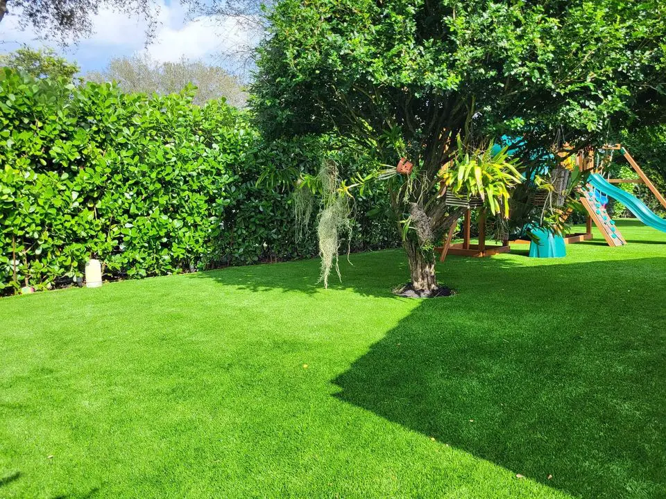 A lush garden with vibrant green grass, possibly enhanced by artificial grass installations, features a leafy tree with hanging plants and a wooden playground set complete with a slide. Bathed in sunlight under the clear blue Deer Valley, AZ sky, this scene is perfect for pets to enjoy on pet turf.