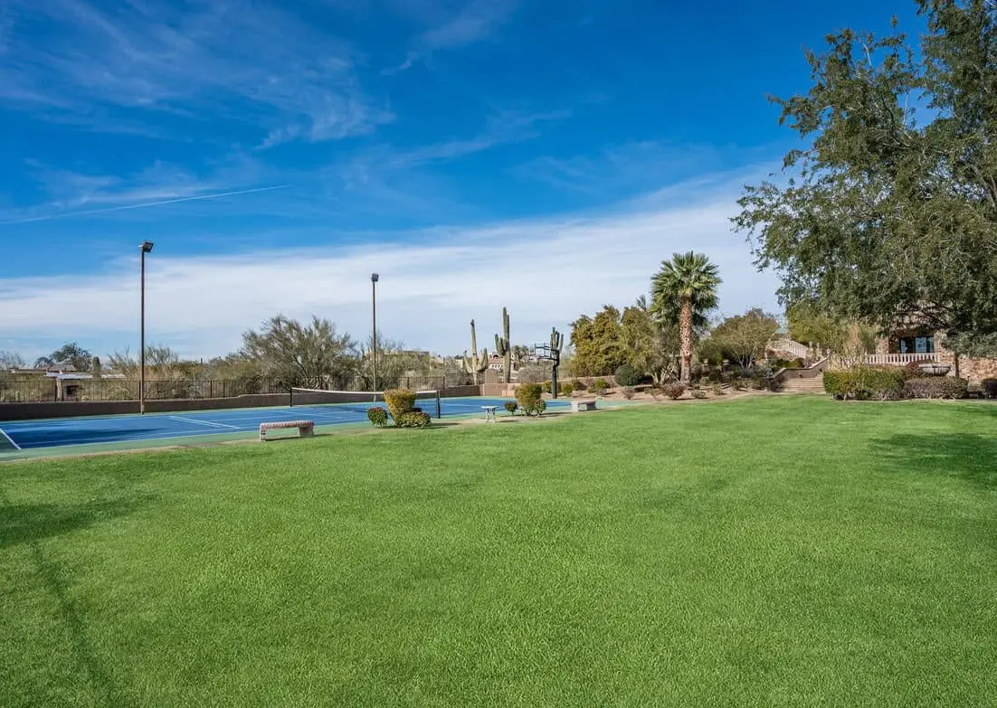 A lush green lawn of artificial grass under a clear blue sky, bordered by desert shrubs and cacti. To the left, a tennis court with a net and surrounding fence is visible. A few scattered palm trees and a building in Deer Valley are in the background.