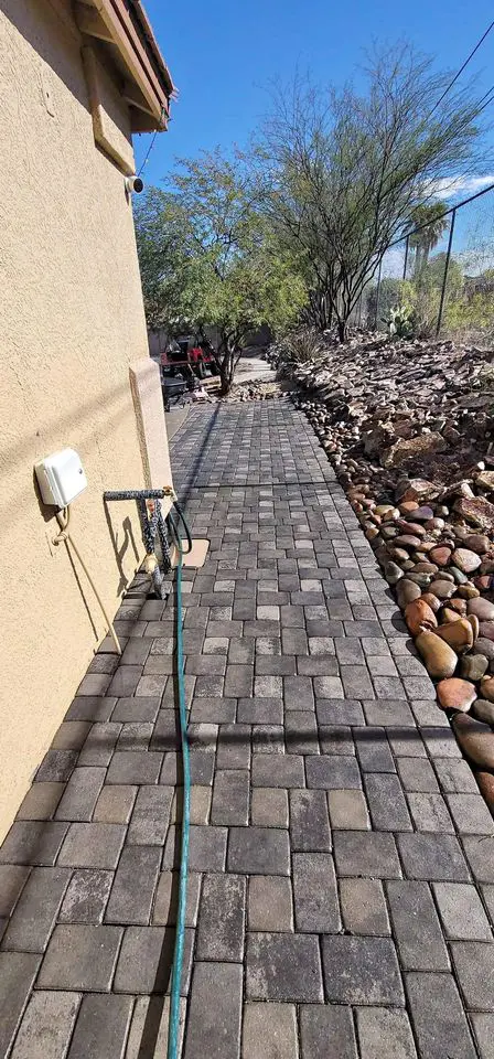 A paved walkway beside a beige building in Deer Valley, AZ, showcases impressive curb appeal, surrounded by desert landscaping with rocks and sparse trees. Paver installations complement the scene as a green hose stretches along the path under a clear blue sky.