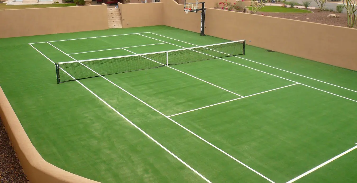 A small green sports court in North Phoenix, AZ, with white tennis lines and a net at its center. Featuring artificial grass, it includes a basketball hoop at one end. Beige walls and steps frame the area, while desert landscaping provides a scenic backdrop around the court.