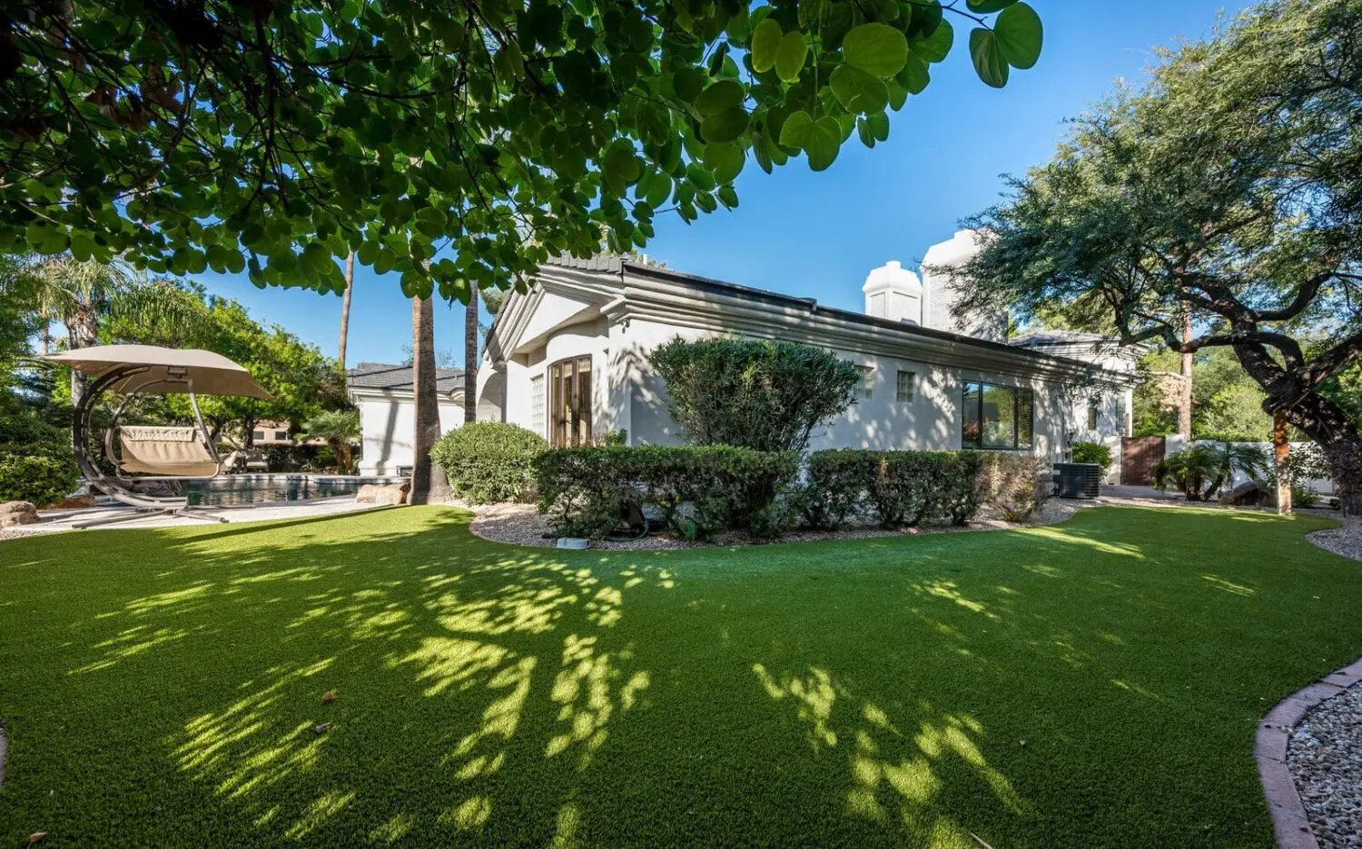 A serene backyard in Deer Valley, AZ, features a well-maintained lawn with a touch of artificial grass installations bordered by bushes and trees. A small pool is visible in the background with a lounge area under a canopy. The sky is clear, and sunlight filters through the leafy trees.