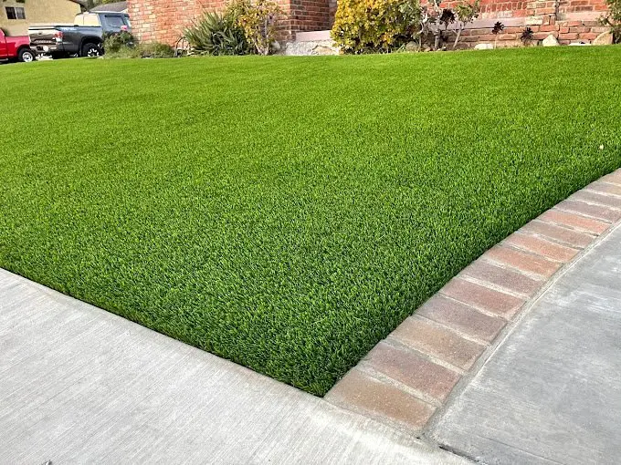 A neatly manicured, vibrant green lawn with a smooth, curving edge meets a light gray concrete sidewalk and a row of red bricks. The background features shrubbery and a brick wall of an Arizona house, showcasing the artistry of artificial grass installations in Deer Valley.