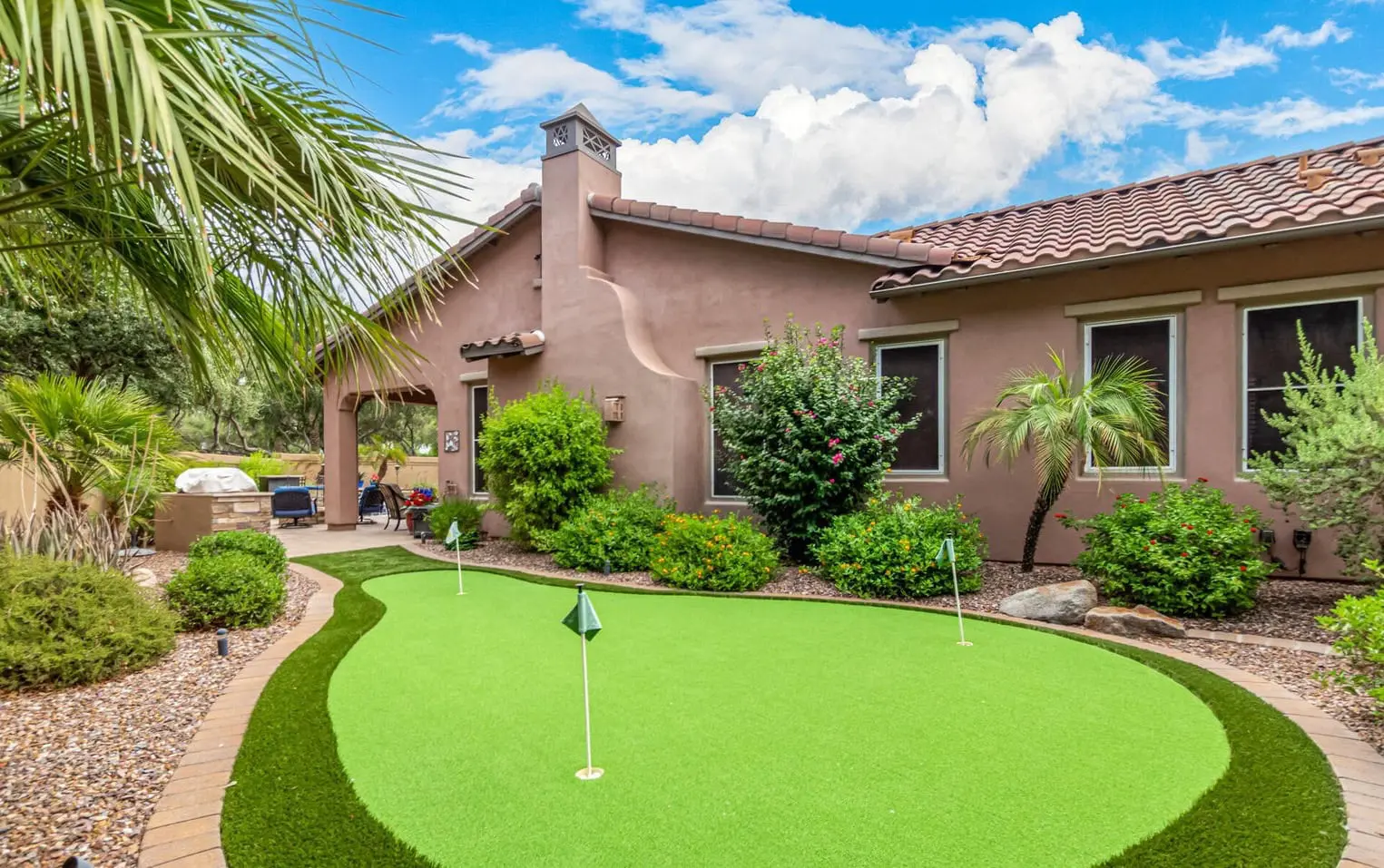 A backyard scene in Deer Valley features a small putting green with lush artificial grass, surrounded by neatly trimmed bushes and palm trees. The house boasts a stucco exterior with red-tiled roofing. The sky is bright with some clouds, creating a sunny and inviting atmosphere.