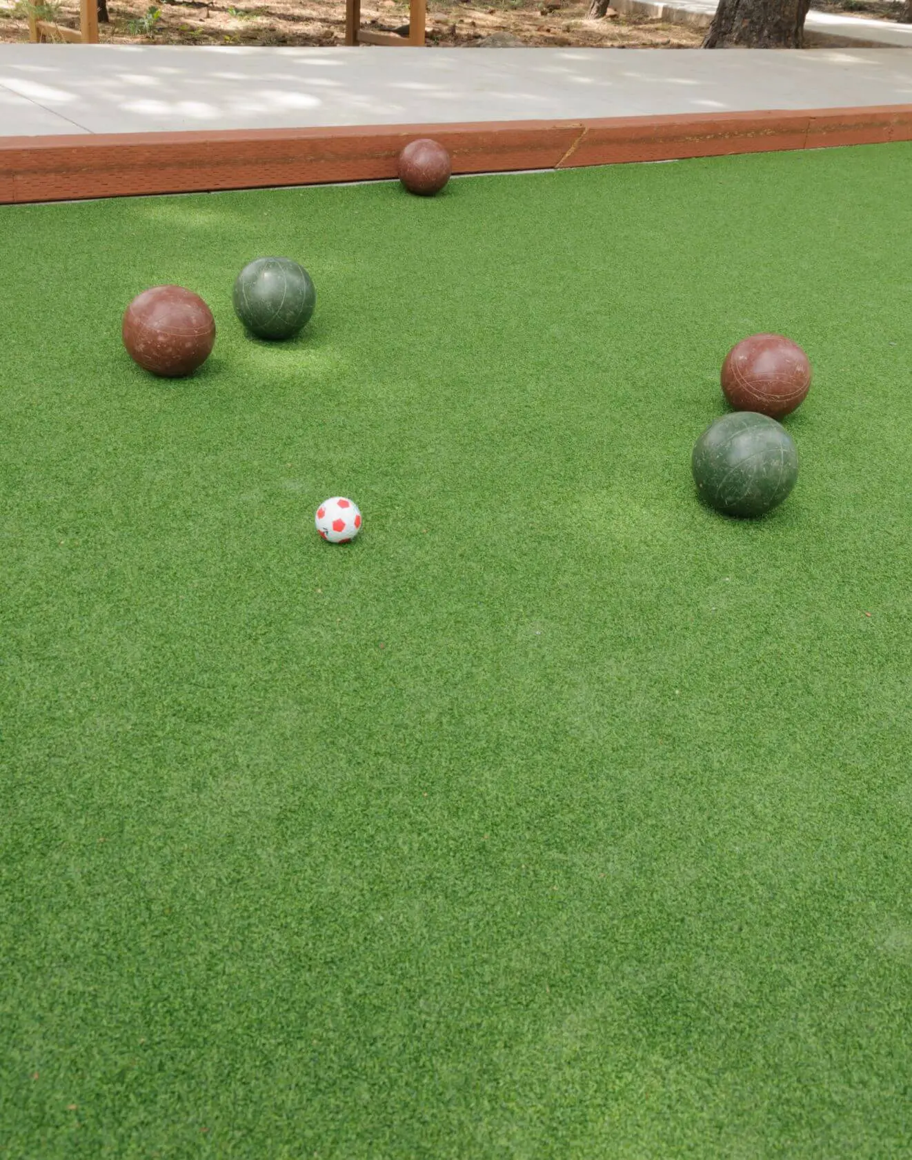 A bocce ball court with four large balls, two green and two brown, scattered on the synthetic turf. A small white ball with red spots is centered among them. The scene features Deer Valley's lush trees and a paved walkway in the background, showcasing top-tier artificial grass solutions.