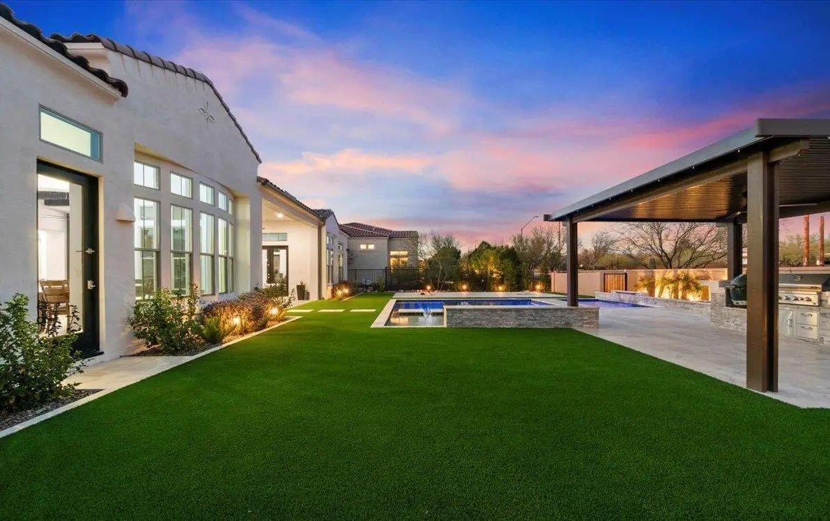 A modern backyard in Arizona boasts a neatly manicured lawn with artificial grass installations, a rectangular swimming pool, and a covered outdoor kitchen area. The vibrant sunset sky suggests the charm of Deer Valley, complemented by the house's large windows and outdoor lighting.