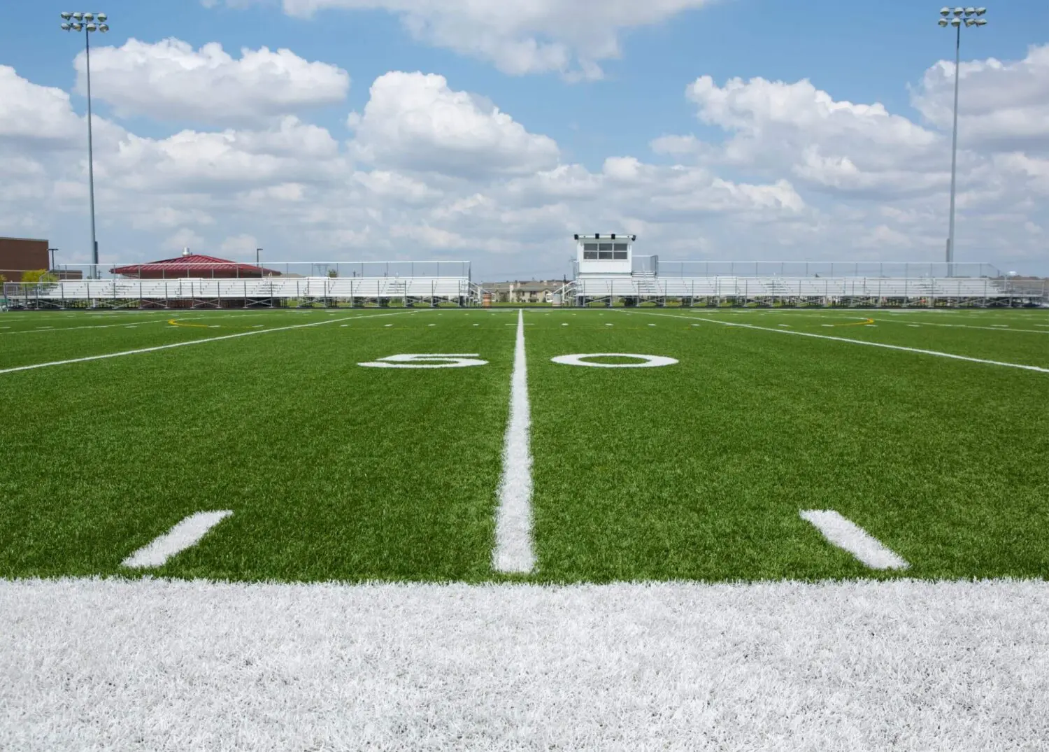 A football field with bright green artificial grass and white yard markings, centered on the 50-yard line. The field is under a blue sky with scattered clouds, with bleachers and a press box visible in the background, perhaps a recent synthetic turf installation in Deer Valley & North Phoenix.