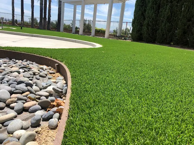 A lush green lawn with neatly trimmed artificial grass installations borders a small rock garden. Smooth, round stones fill the garden area. In the background, white pergolas and tall trees are visible under a partly cloudy sky, reminiscent of serene Deer Valley landscapes.