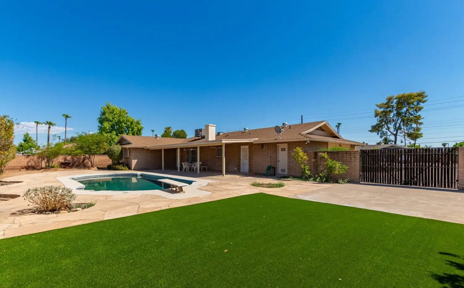 A backyard with a small in-ground pool is surrounded by a stone patio and lush lawns. Adjacent is a green lawn, enhanced by artificial grass installation. A single-story brick house with a covered patio and outdoor furniture sits gracefully under a clear blue sky.