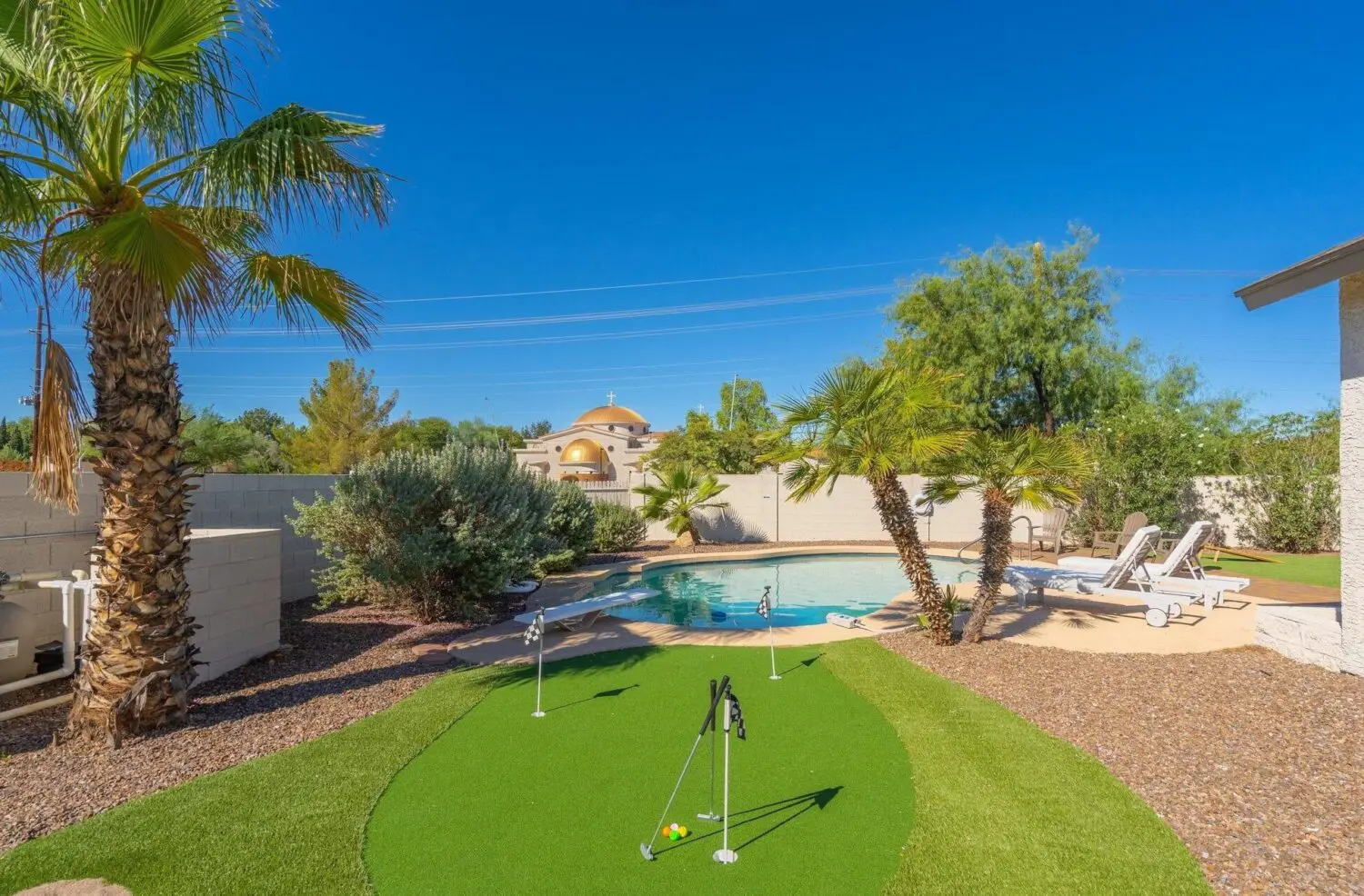 A backyard scene in Glendale AZ with a circular swimming pool surrounded by lounge chairs and palm trees under a clear blue sky. In the foreground, an artificial putting green with golf flags is visible, complemented by expert artificial grass installation and desert landscaping.