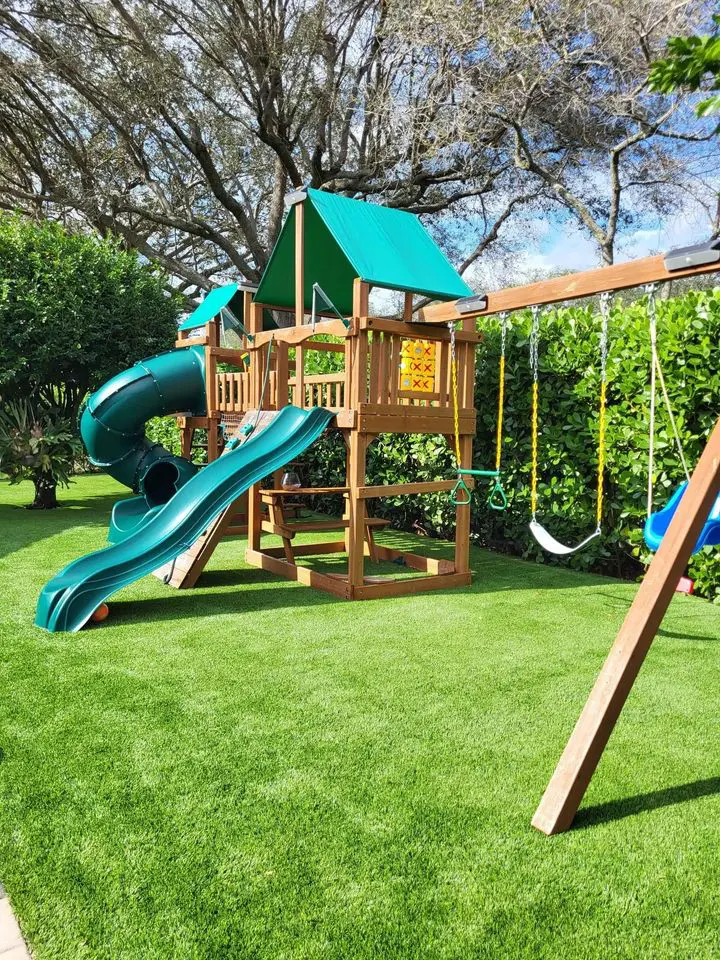 A children's playset on the lush lawns of Glendale, AZ, boasts a wooden structure with a green slide, spiral slide, swings, and a small climbing wall. Tall trees and vibrant greenery envelop the play area under a clear blue sky.