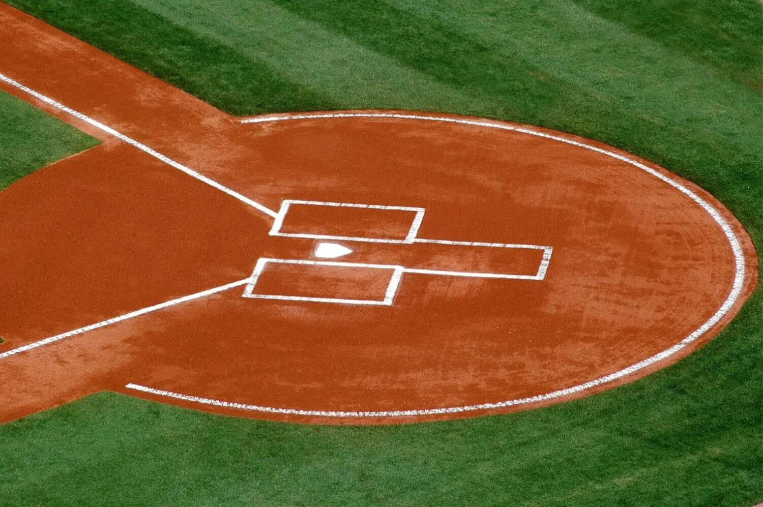 An aerial view of a baseball diamond in Glendale, AZ reveals the vibrant contrast between the reddish-brown dirt around home plate and the lush lawns of bright green outfield grass. White lines mark the batter's boxes, perfectly framing the centered plate.