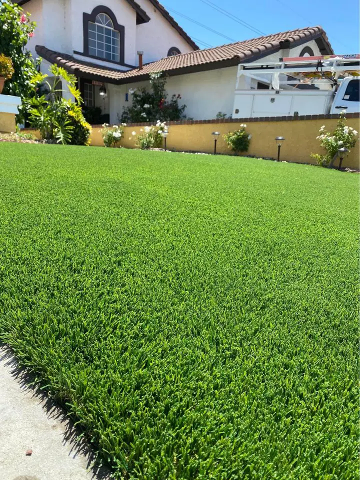 A well-maintained lawn with vibrant artificial grass, bordered by colorful flowers and shrubs. In the background, a two-story house with a tiled roof and a white vehicle parked in the driveway, showcasing low-maintenance turf perfect for Phoenix, AZ's climate.