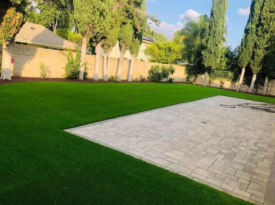 A backyard in Sun City West features a neatly trimmed green lawn and a large paved patio area. Tall trees and a brick wall border the yard, with synthetic turf enhancing the space. A garden hose is coiled on the patio, adding to its charm.