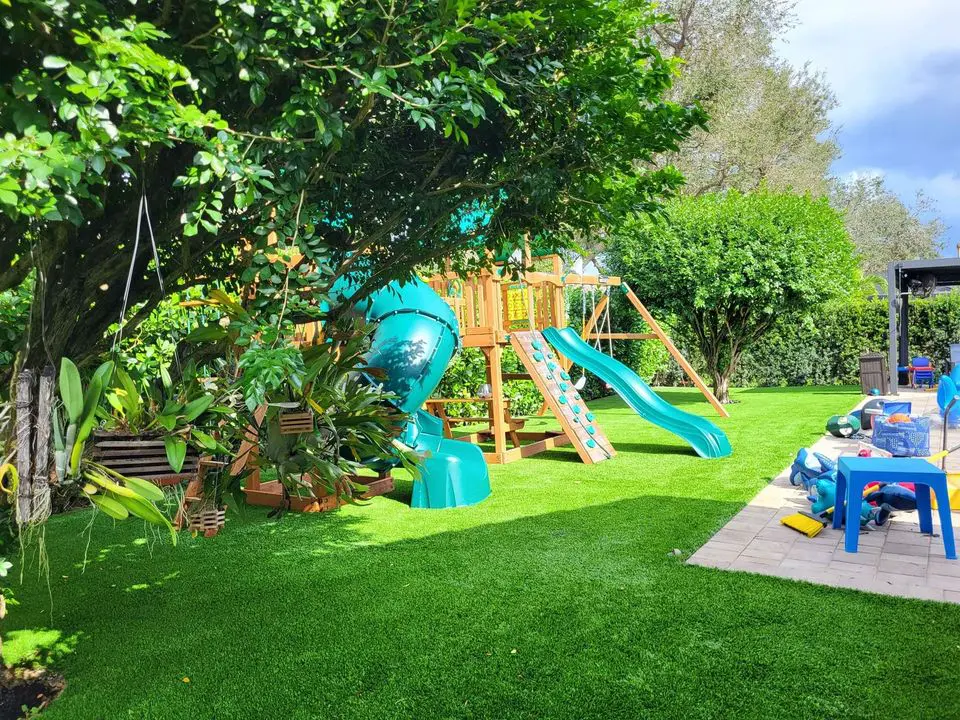 A backyard playground in Sun City West boasts synthetic turf with vibrant green hues, featuring a wooden playset with a blue spiral slide and a traditional slide. The area is surrounded by leafy trees and shrubs. Nearby, scattered toys and a blue plastic chair adorn the patio.