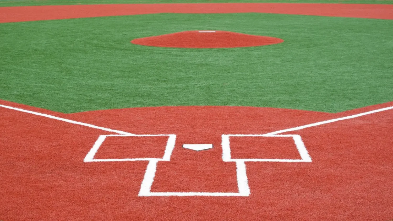 A baseball field with vibrant red synthetic turf and white lines marking the batter's boxes, home plate, and the pitcher's mound. The green infield contrasts beautifully with the red. The view is from behind home plate toward the pitcher's mound in Sun City West.