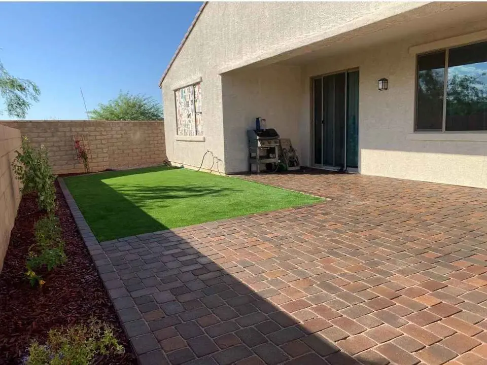 A backyard in Sun City West features a brick patio and a small synthetic turf area. A covered patio with a sliding glass door is attached to the house. Along the fence, small plants thrive in a flower bed, while a barbecue grill is conveniently near the patio door.