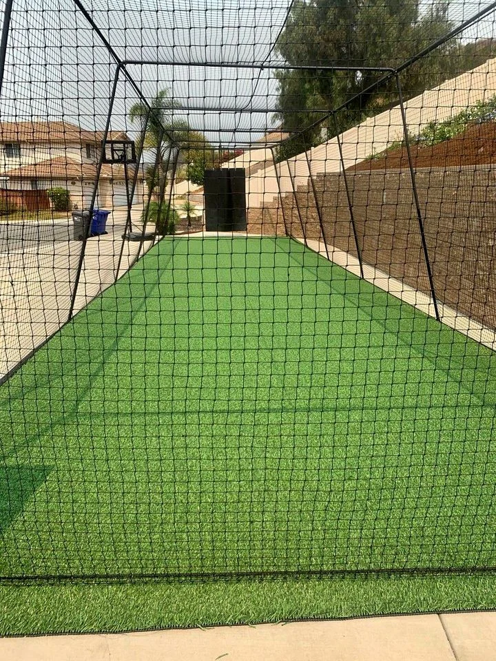 A backyard sports setup in North Phoenix features a long stretch of synthetic turf enclosed by a black netted cage for practicing. The area, surrounded by stone walls and trees, also boasts a basketball hoop visible in the background.