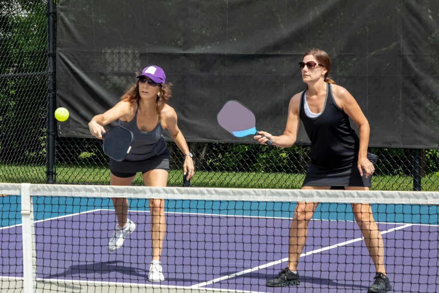 Two women playing pickleball on a court with synthetic turf in Deer Valley. Both are wearing athletic clothing; one sports a purple cap, and the other wears sunglasses. Positioned to hit a yellow ball, they hold paddles in front of a black net against the vibrant artificial grass backdrop.